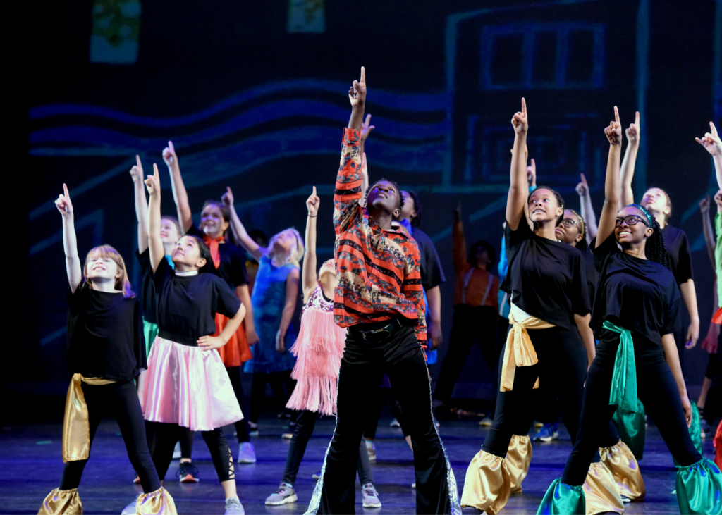 A performance shot of middle school aged kids in colorful costumes all pointing one hand towards the sky.