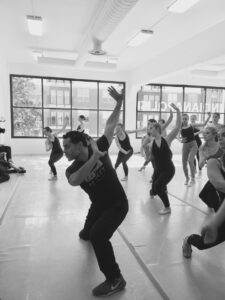 Black and white photo of Joshua Bergasse in a jazz position leading a group of dancers in a window-filled studio.