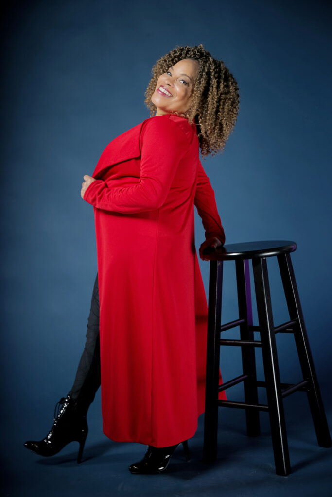 Debbie Wilson wears heeled boots and a long red coat and poses with her hand on a tall stool.
