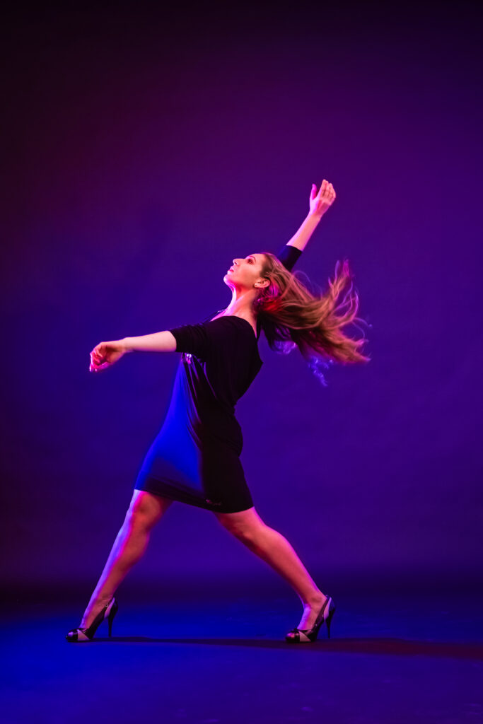 Emily Bufferd poses in front of a deep blue-violet backdrop in a dark velvet dress, her long hair flying behind her as she twists her body to the right and lifts her arms.