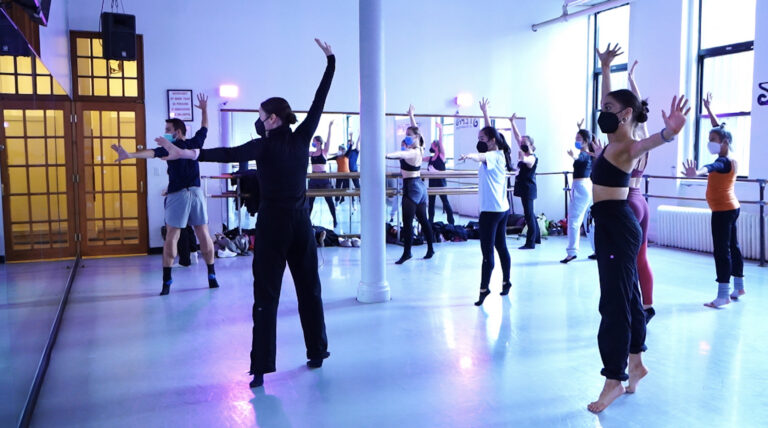 Emily Bufferd teaching at Steps on Broadway. Bufferd wears all-black athletic clothing and demonstrates a combination at the front of a purple-lit classroom. Her students mirror her movements.