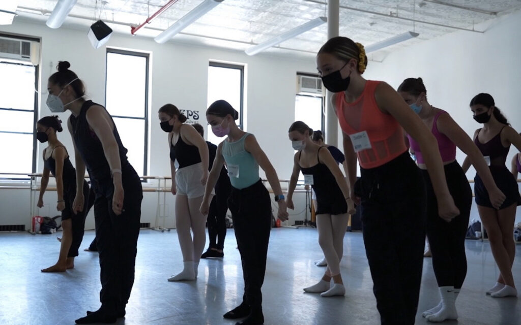 Emily Bufferd teaching the Steps Youth Program in 2022. Bufferd wears black athletic clothing as she leads a class of young female dancers; they all wear masks and lean forward, their arms at their sides.