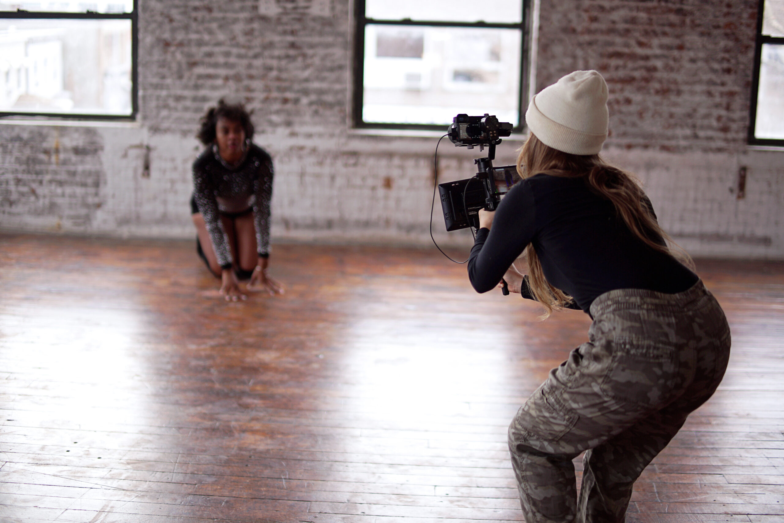 Woman leans over, filming a dancer kneeling on the ground in a brick-lined studio.