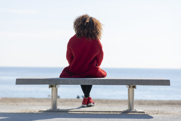 Rear view of a woman in a red coat and shoes sitting on a bench, looking out at the ocean.