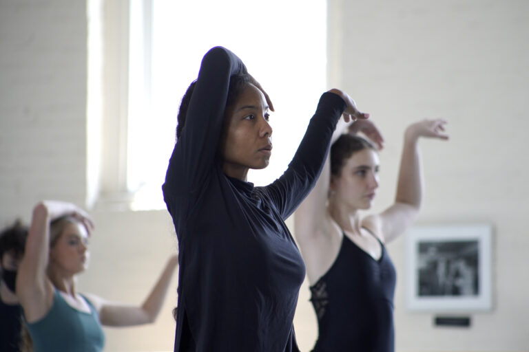 Sidra Bell stands in a room of students copying her movements. She has one hand on her head and the other arm extended.