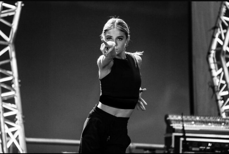 A black and white photo of Hailey Bills shows her quads-up. She dances in a black crop top and black pants, her long blonde hair in a low ponytail. She points front intently with her right arm, twisting her torso slightly away, pulling her left arm back by her waist.