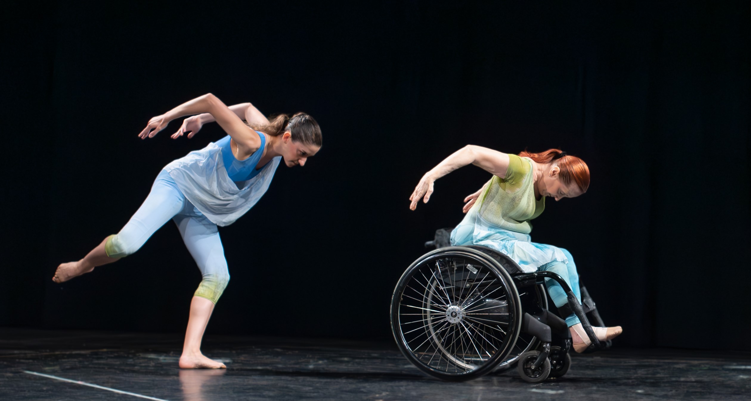 Performance shot of a dancer in blue and a dancer in green, who's in a wheelchair, doing the same movement with arms behind their backs and elbows bent.