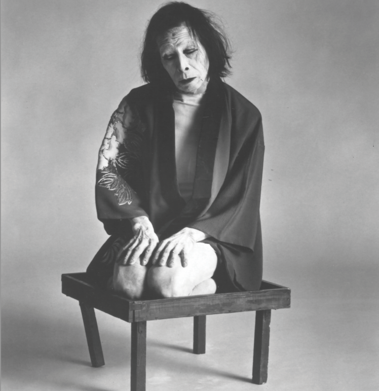 A black and white photograph of Kazuo Ohno, dressed in only a silk robe, kneeling on a low wooden stool.