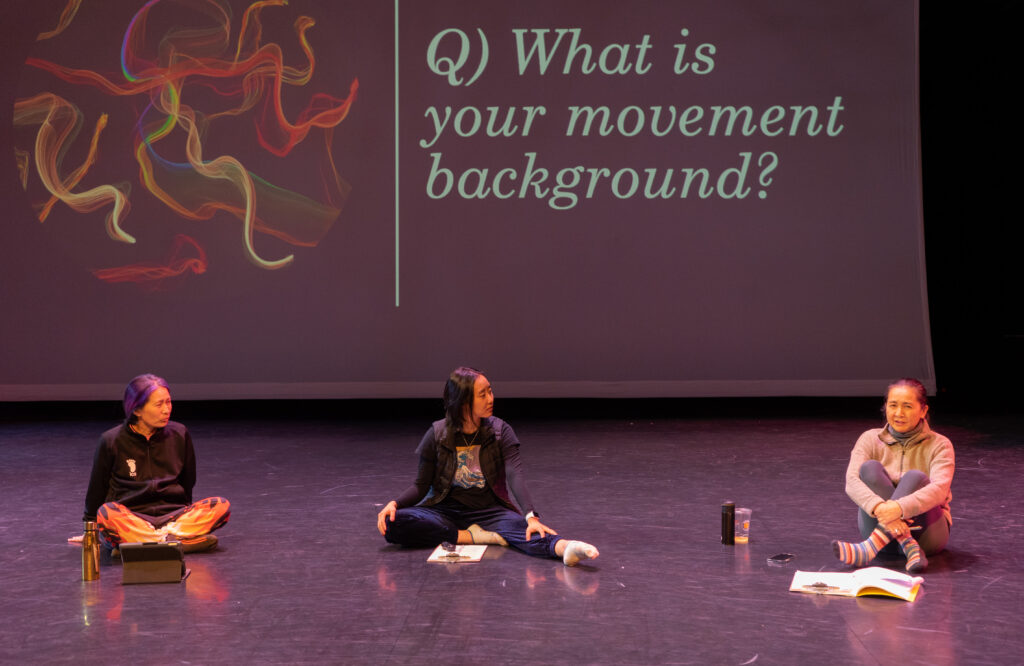 Three Asian dancers sit on the floor of a stage talking with a projected screen behind them. 