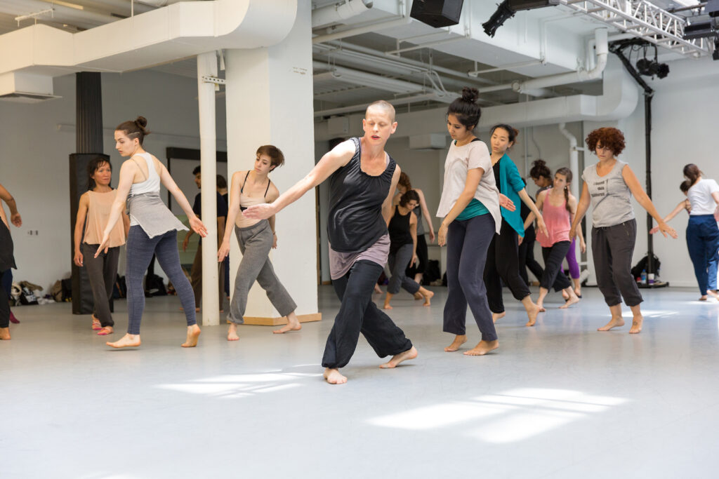 Class photo with Jennifer Nugent center of the studio, wearing a blank tunic and black loose pants in a twisted position.
