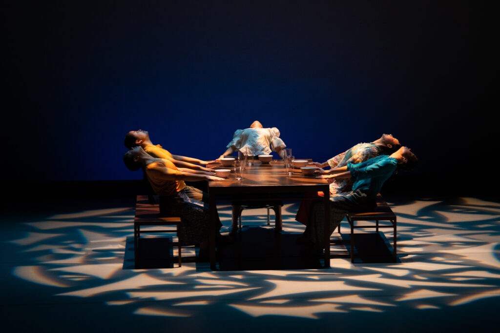Performance image of five dancers sitting at a large wooden table, leaning all the way back so they're looking at the ceiling. 