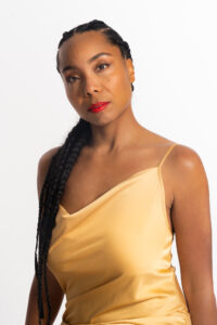 Sidra Bell poses in a yellow top and red lipstick with her hair over one shoulder.