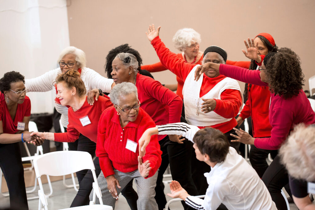 Haas, in a white top, leads a group of older Black dancers wearing red tops in movement.