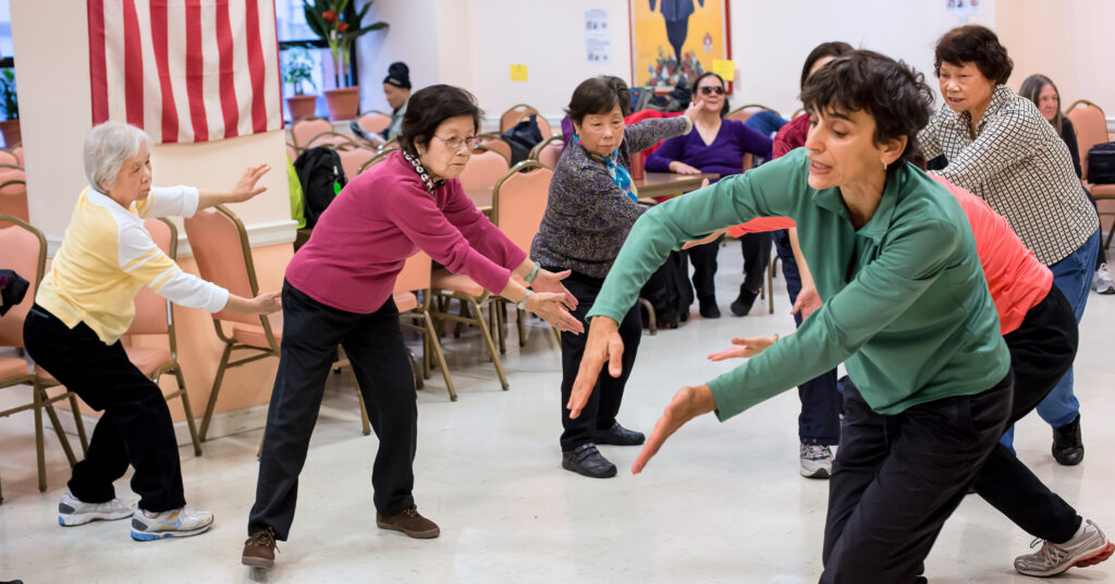 Naomi Goldberg Haas, wearing a green top and pants, leads a group of senior Asian women through a movement exercise, guiding with her hands.