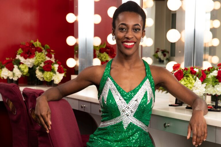 Alexis Payton wears a sparkly green and white Rockettes costume and sits in front of a dressing room mirror.