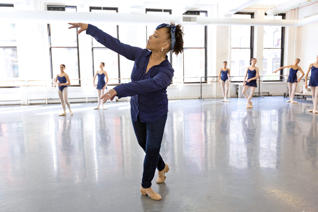 Studio shot of Caridad Martinez, wearing a blue shirt, pants, and dance shoes, standing in a turned out position with her right leg behind her, gesturing with her arms and looking towards them.