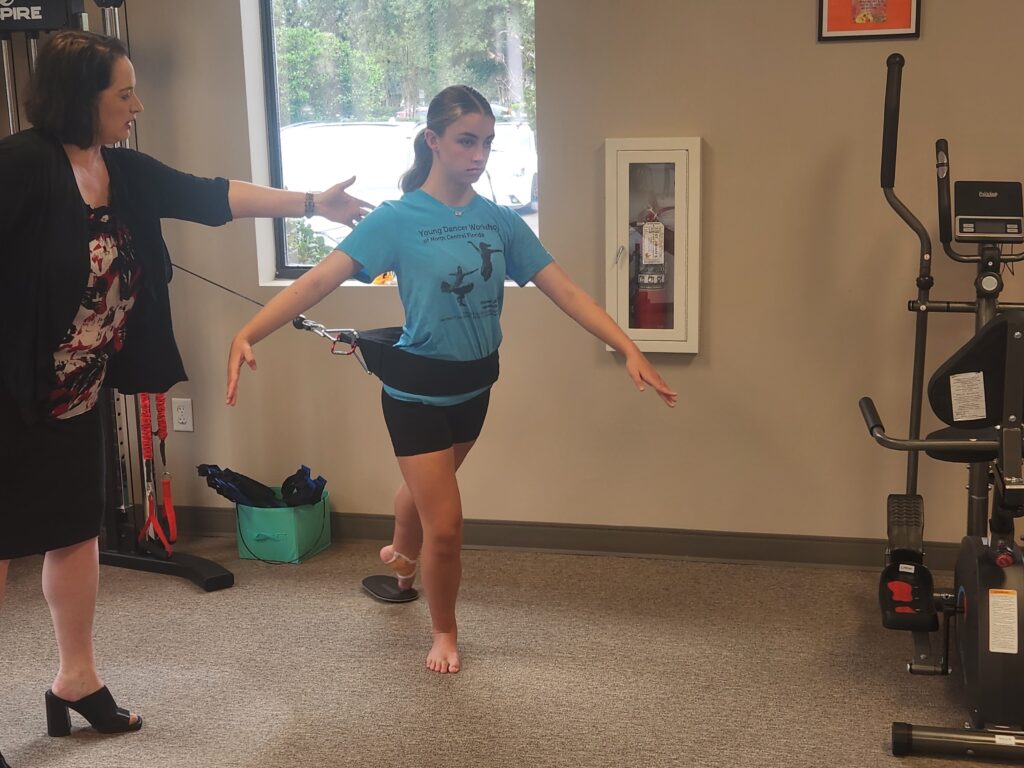 Emily Becker in a PT studio, adjusting a dancer standing in a low arabesque with resistance bands.