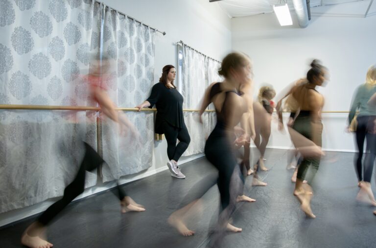 Image is focused on Lauren Setten, who leans against a ballet barre watching dancers who are blurred.