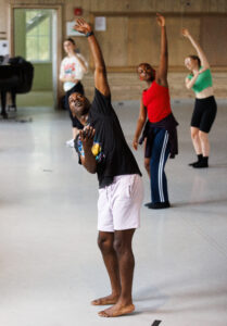 Russell teaching in the studio with his knees bent and left arm in the air, wearing a black t shirt and white shorts.