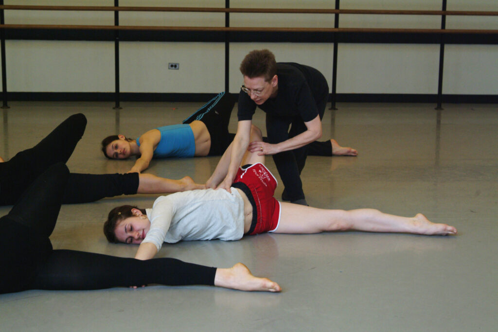 Irene Dowd bends forward to guide a dancer as they perform Dowd's "Spirals" sequence. The dancer lies on the floor, face down. One leg bends and hooks behind them, opening up their hip; their opposite arm reaches long on the floor away from the bending leg.