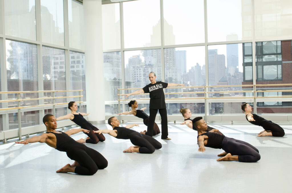 Ana Marie Forsythe, wearing all black, guides a group of dancers dressed in black who are on their knees leaning back in a flat back with arms outstretched.