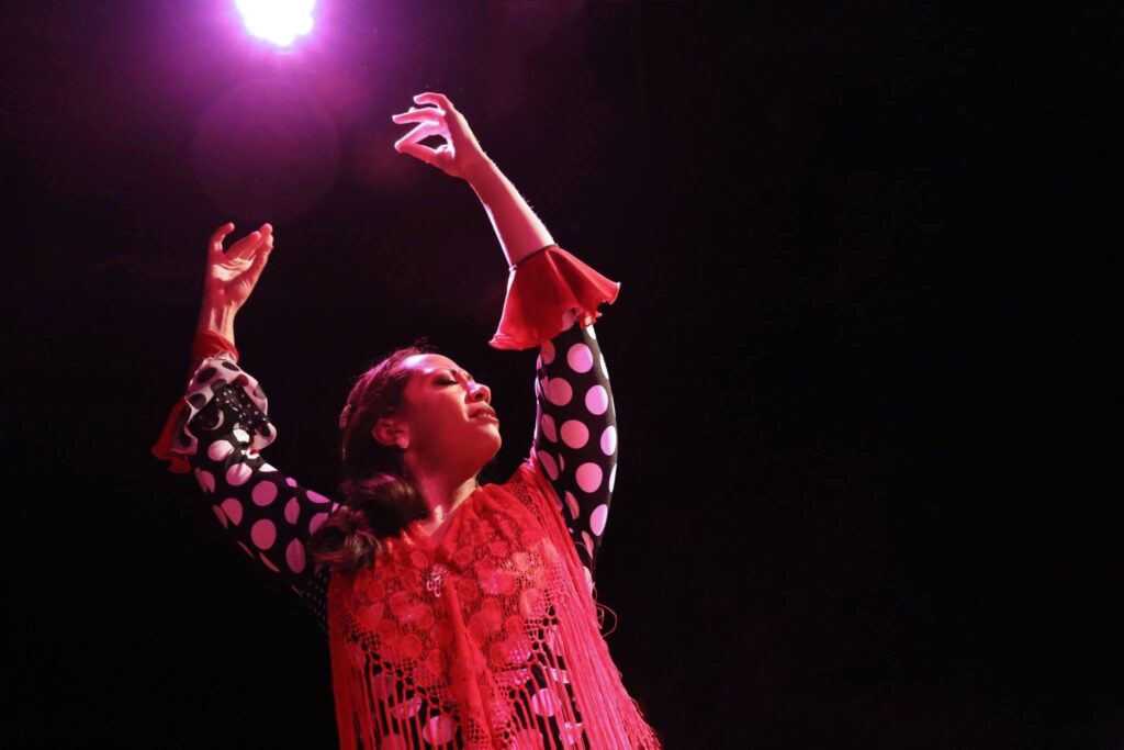 Performance photo of Xianix Barrera in black and white polka dots and a red shawl with her hands in the air, lit against a dark background.