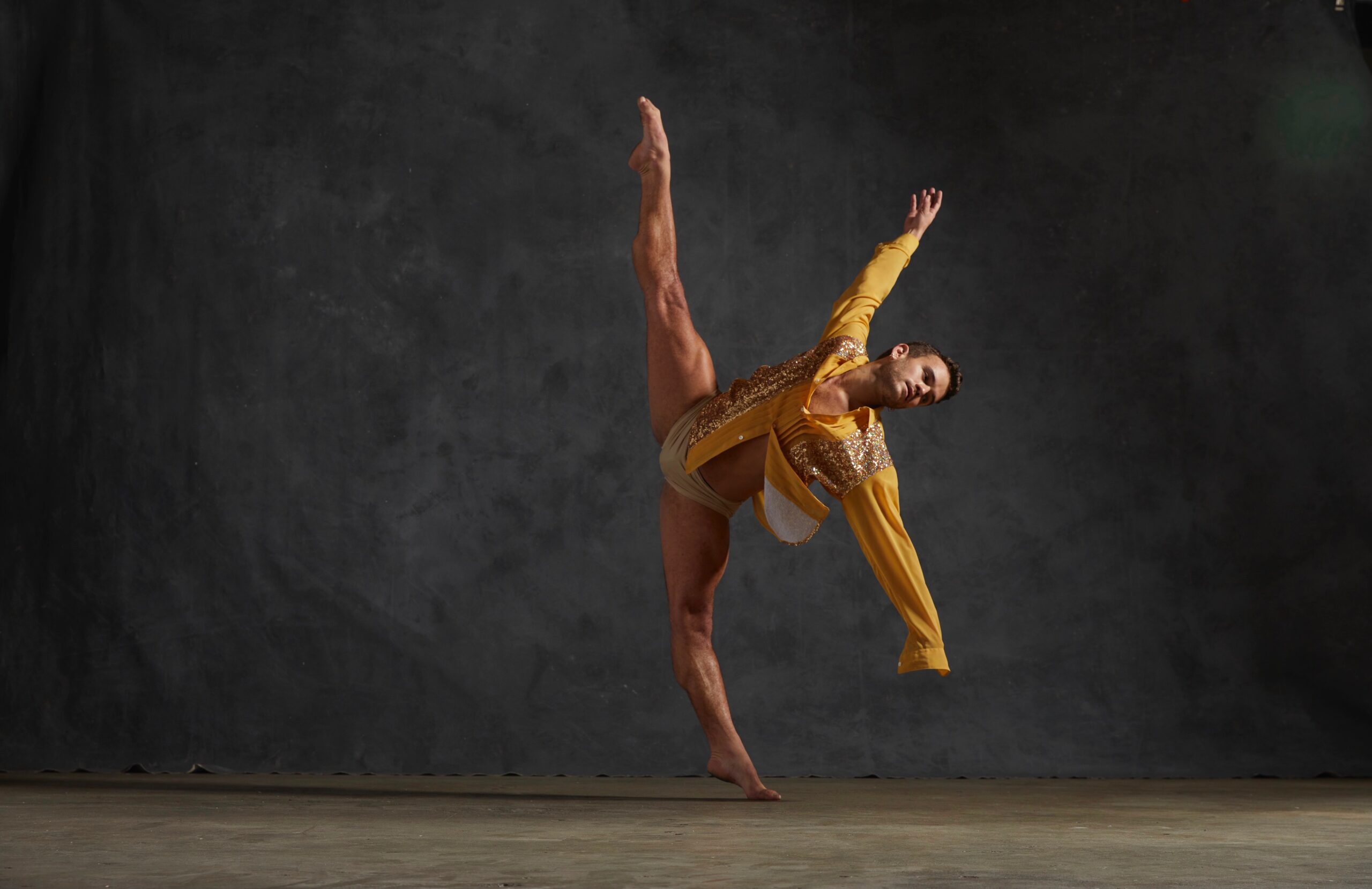Dance Reed Tankersley in a dance pose with legs in a sideways split and arms outstretched. He wears an embellished yellow costume.
