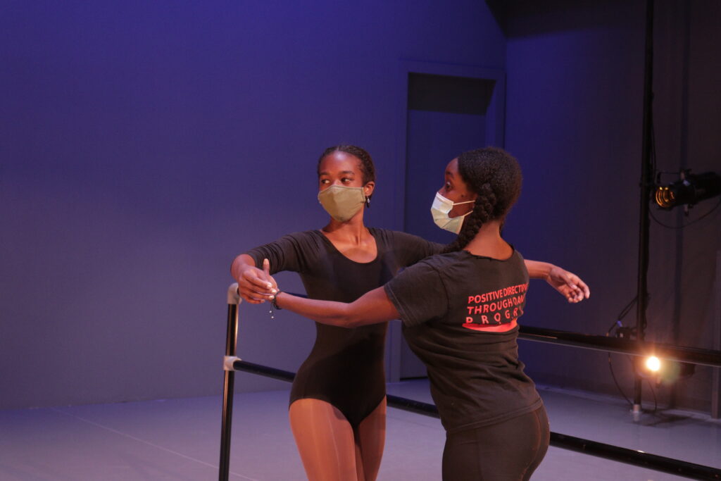 Masked dance teacher from Dance institute of Washington assisting a masked student at the barre.