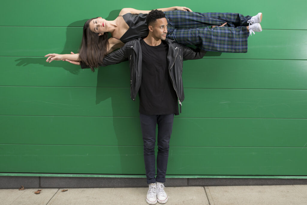 Dancer Chris Grant stretching arms to the side with his partner Lauren Grant lying horizontally across. They are against a green background.