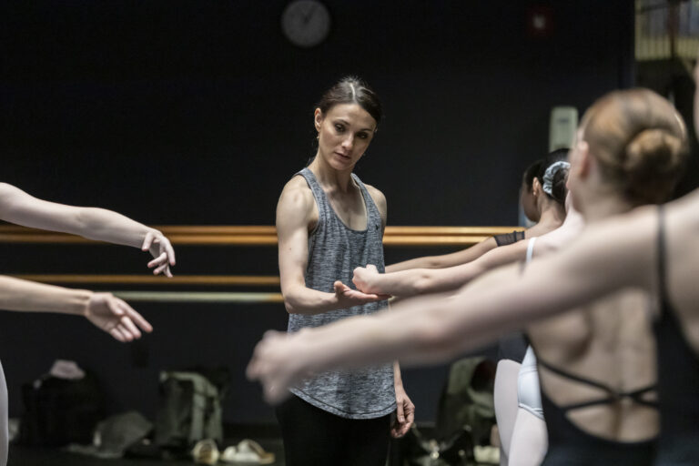 Sarah Lane gently places a hand under a dancer's wrist in second position. She looks thoughtful, watching blurry dancers in the foreground working at barre.