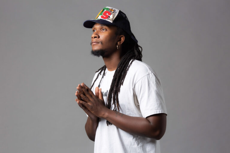 Headshot of dance Saleh Simpson against a grey background. He wears a baseball cap and his hands are joined together.