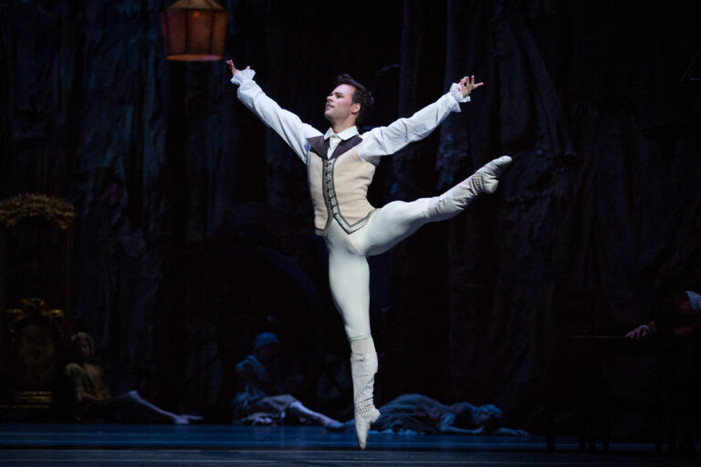 Alexander Campbell jumps with one leg extended side, just above 90 degrees. His arms are slightly above chest height as they extend to the side, palms up. He is costumed in a cream colored vest over a white shirt and matching tights and ballet slippers.