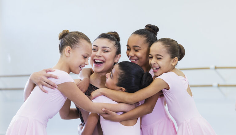 A multi-ethnic group of girls hugging their dance instructor, in a dance studio, smiling.