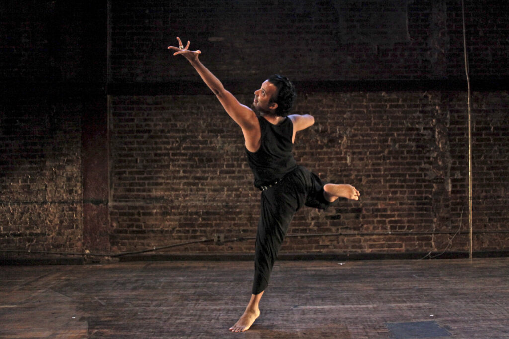 Hari Kirshnan dances in a room with an exposed brick wall. His back is to the camera as he balances on the ball of one foot, looking over his shoulder as he extends a splayed hand up to the corner. His other hand is pressed to his chest; his working leg kicks up and back in almost a flexed foot back attitude.