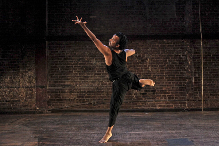 Hari Kirshnan dances in a room with an exposed brick wall. His back is to the camera as he balances on the ball of one foot, looking over his shoulder as he extends a splayed hand up to the corner. His other hand is pressed to his chest; his working leg kicks up and back in almost a flexed foot back attitude.