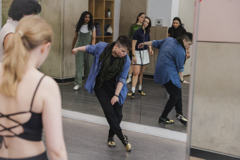 Dre Torres demonstrates for a small class of tap students. She is a young white woman with her hair styled in an undercut; she wears a denim button down open over dark pants, topped with a dark green, drapey scarf. She faces her students as she crosses one foot in front of the other, head dropped toward her working leg in a jazzy style.