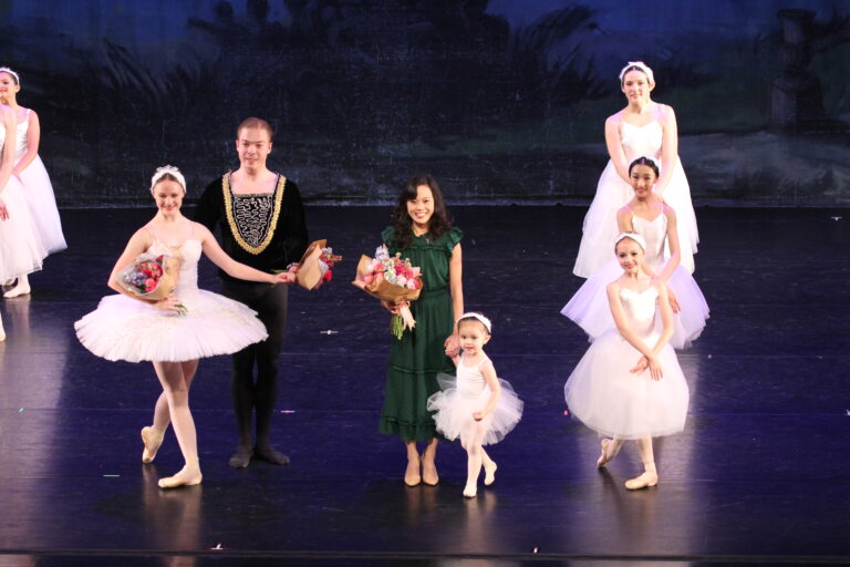 Misa Oga wearing a green dress and holding a young ballerina's hand among other dancers on stage.
