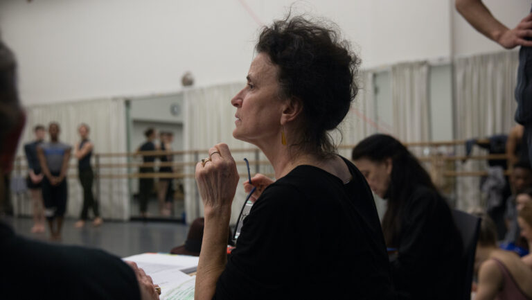 Terese Capucilli is shown in profile from the chest up, sitting at the front of a dance studio. She wears a dark shirt and holds a pair of eyeglasses in her right hand as she draws her other hand up towards her chin. She wears her dark curly hair piled on her head. In the background, slightly blurred, are dancers taking a pose, while other dancers and rehearsal assistants sit alongside Capucilli.