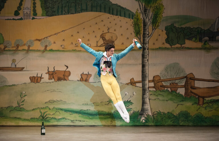 Iain Mackay in performance. He glances down toward his feet mid-assemblé, arms jauntily raised to his sides. In one hand is a bottle of wine. He is brightly costumed in pastel yellow tights and a teal frock coat over a patterned shirt. The backdrop is pastoral.