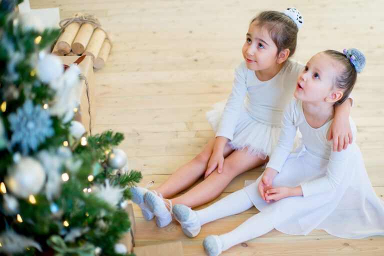 Two young ballet dancer sitting near Christmas tree on the wooden floor and smile