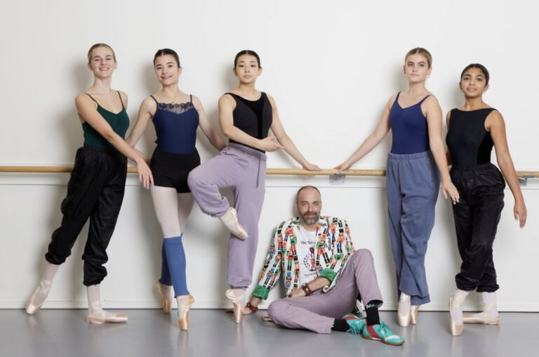 Adam Holms sitting on the floor while his students surround him standing at the ballet barre.