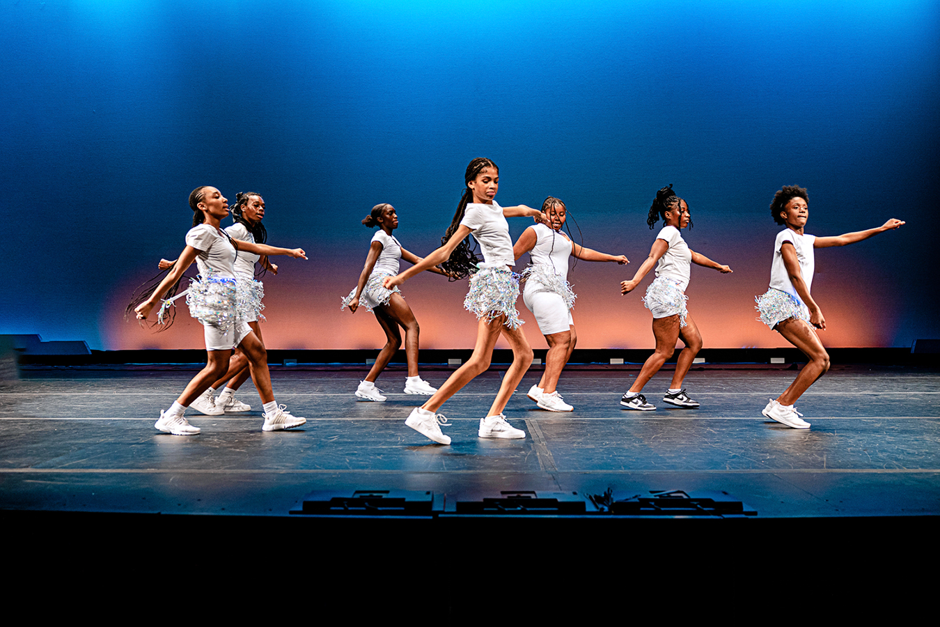 Young girls performing on stage in silvery/white costumes against a blue background.