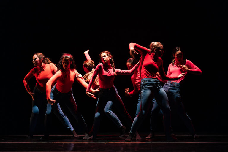 Seven dancers costumed in red turtlenecks and blue jeans perform on a darkened stage. They create angular, asymetrical shapes using negative space, some connecting with hands on each other's sides or arms.