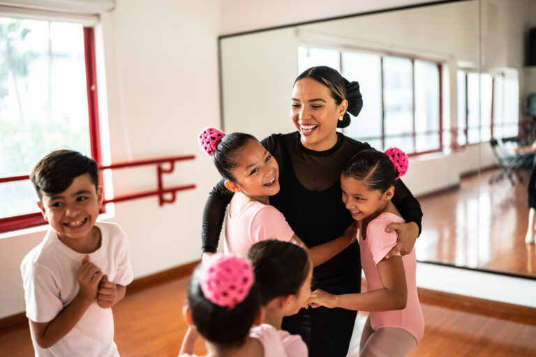 A smiling brunette ballet teacher wearing black is hugged by a group of students. The girls wear pink leotards and the boy wears a white t-shirt.