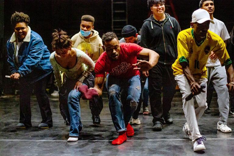 A black dancer leads a diverse group of students in a low, grounded movement.