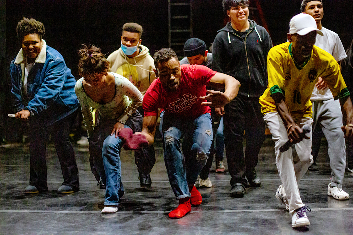 A black dancer leads a diverse group of students in a low, grounded movement.