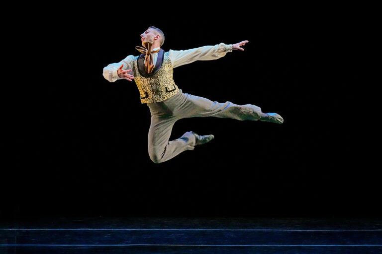 Richard Villaverde wears a long-sleeved top, vest, and pants, leaping through the air against a black background.