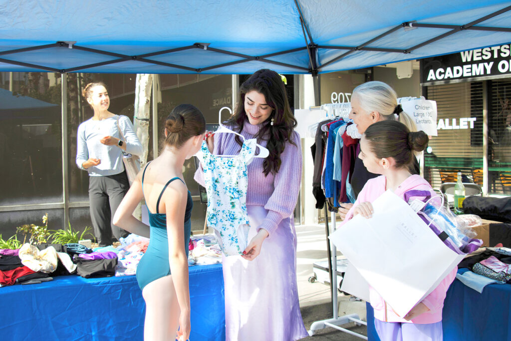 A brunette woman shows a young dancer a leotard, while another dancer and an older teacher look on. The scene is outside.