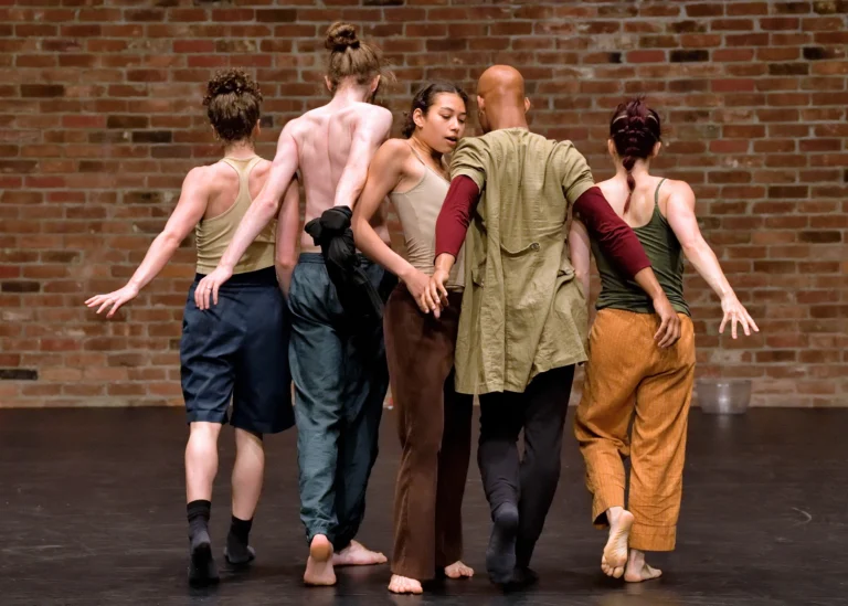 A group of five individuals. Two on the left and two on the right are facing the brick back wall and appear to be walking towards it. The figure in the middle is sideways.