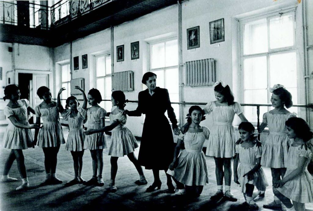 A black and white image of a dance teacher in a long black dress with young students in old-fashioned dancewear.